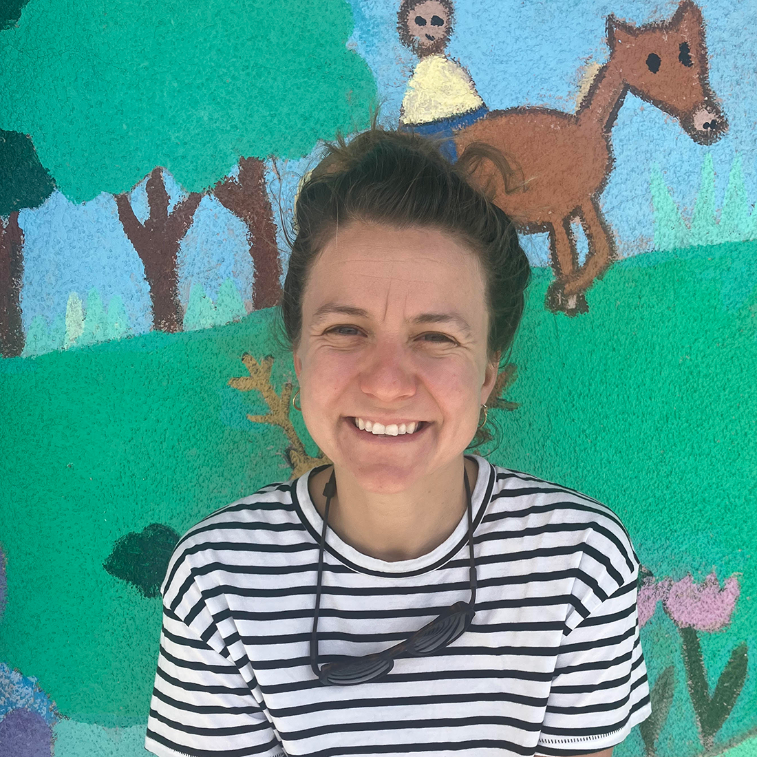 zoe pantera is a caucasian woman with short brown hair. she is wearing a black and white striped tshirt and is posed in front of a mural featuring a childlike drawing ofhorse and rider
